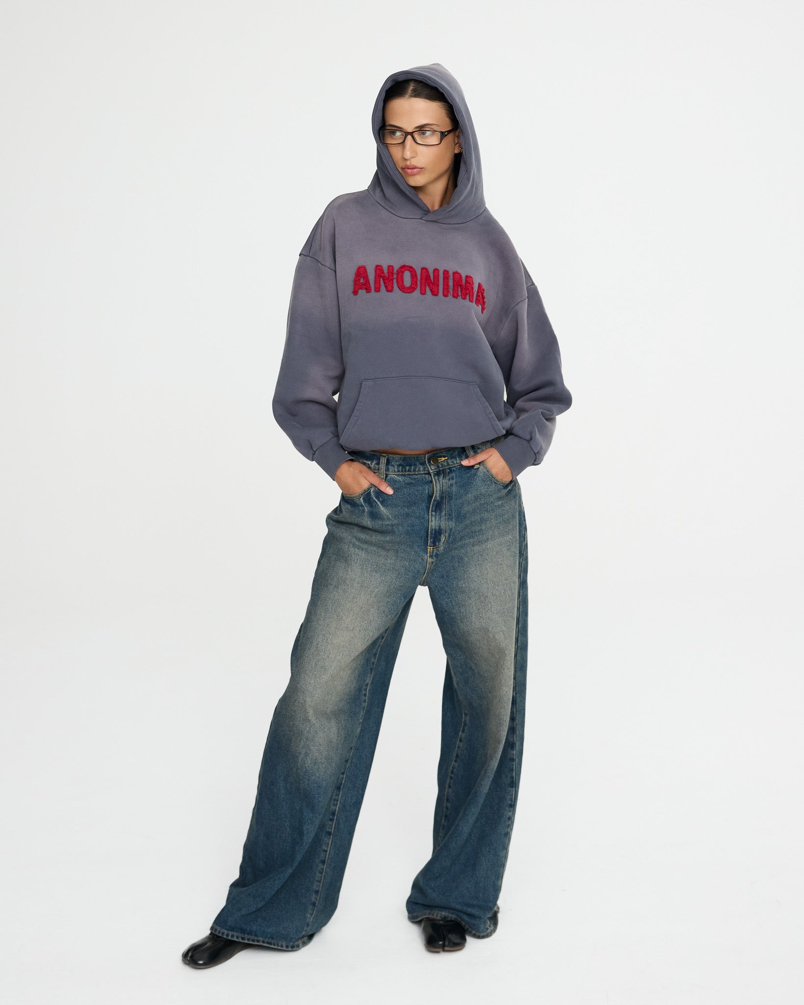Person wearing a gray hoodie with 'ANONIMA' text and blue jeans on a white background