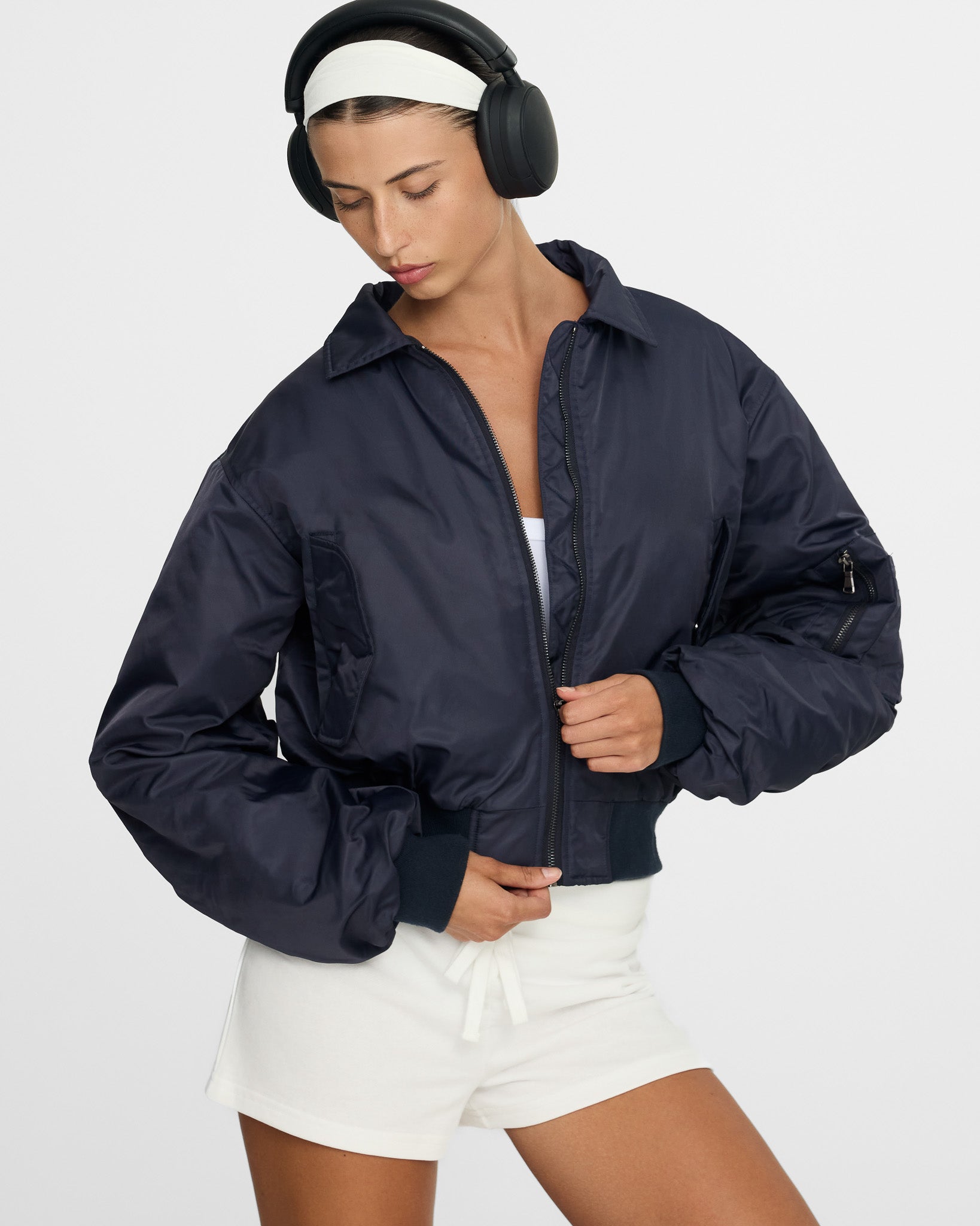 Person wearing a navy jacket and white shorts with headphones on a light gray background