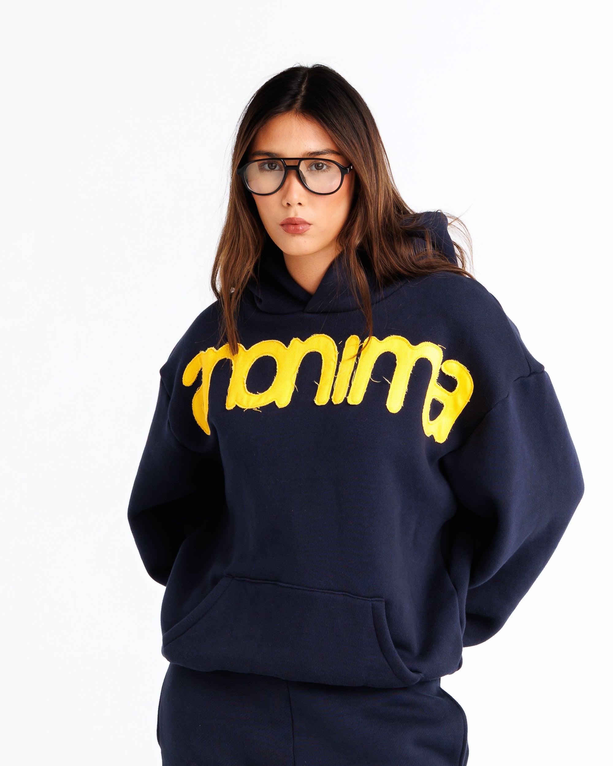A woman wearing a navy blue oversized hoodie with a bold yellow 'Anonima' graphic across the chest, paired with matching navy pants, photographed against a plain white background.