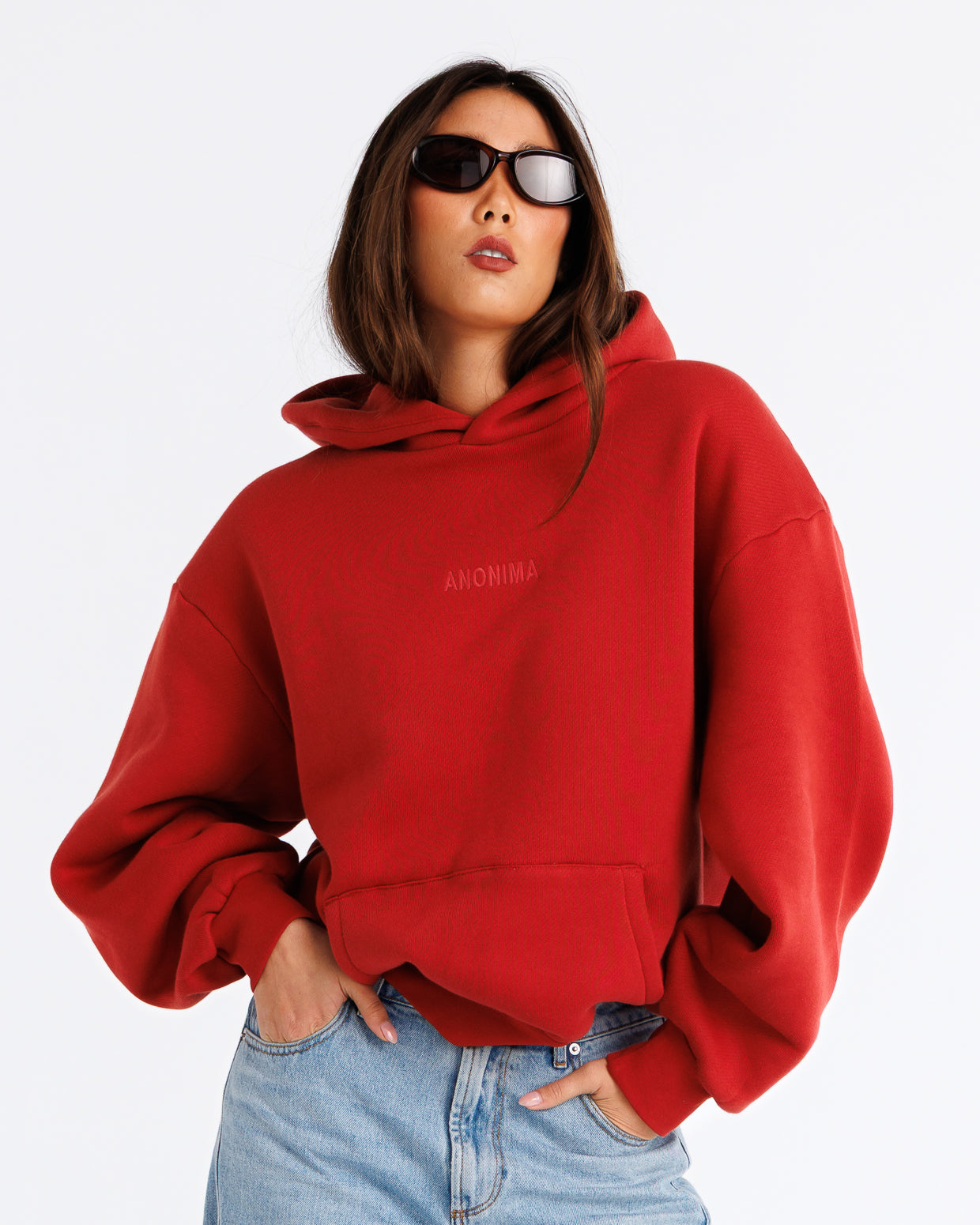 A woman wearing a bright red oversized hoodie with 'ANONIMA' printed in tonal red on the chest, paired with light blue jeans and black sunglasses, against a plain white background.