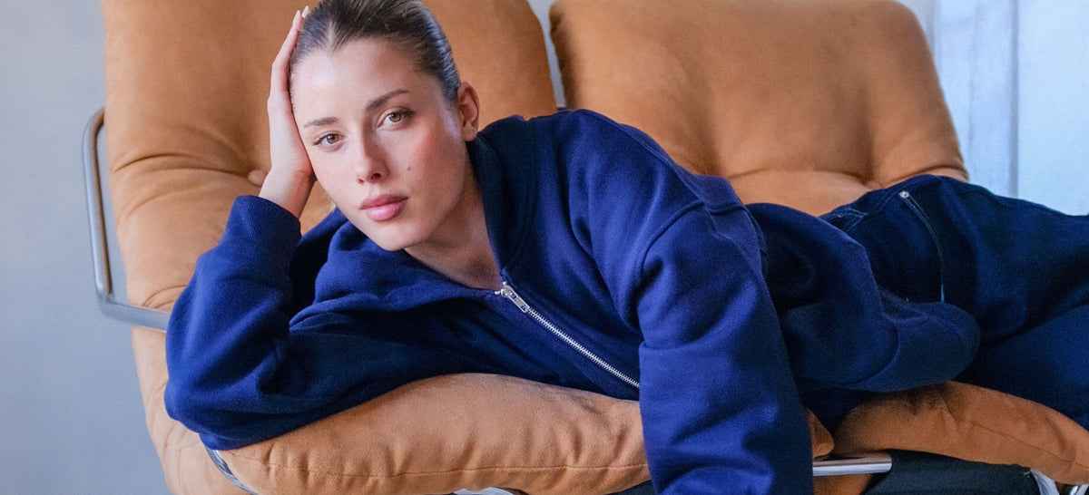A woman wearing a navy blue zip-up hoodie and matching sweatpants reclines on a tan leather lounge chair in a modern room.