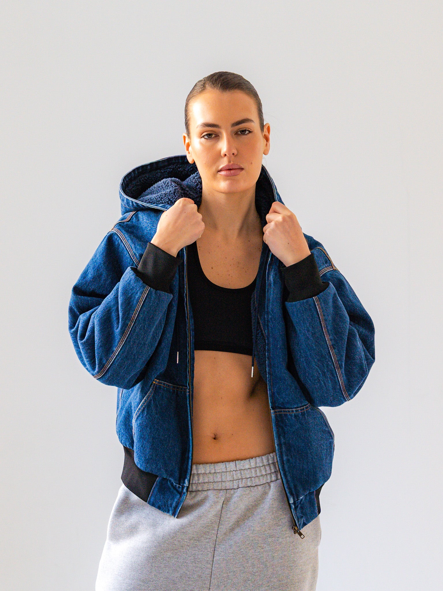 Person wearing a blue denim jacket over a black crop top and gray sweatpants on a light gray background